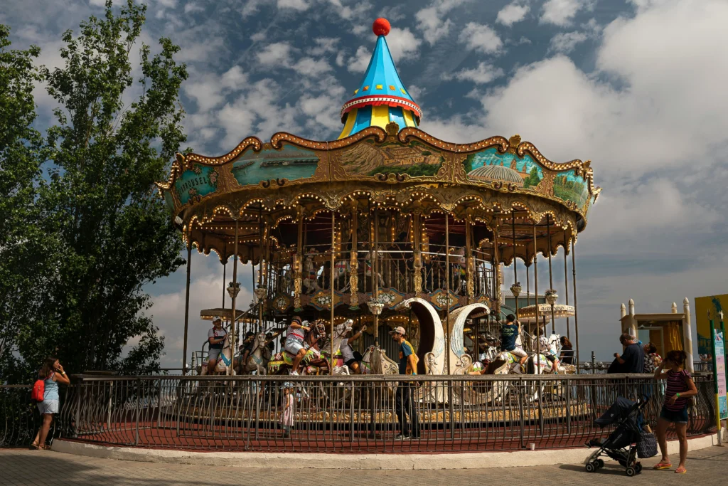 Colorful two-story carousel with ornate decorations and horse figures, surrounded by people on a partly cloudy day.