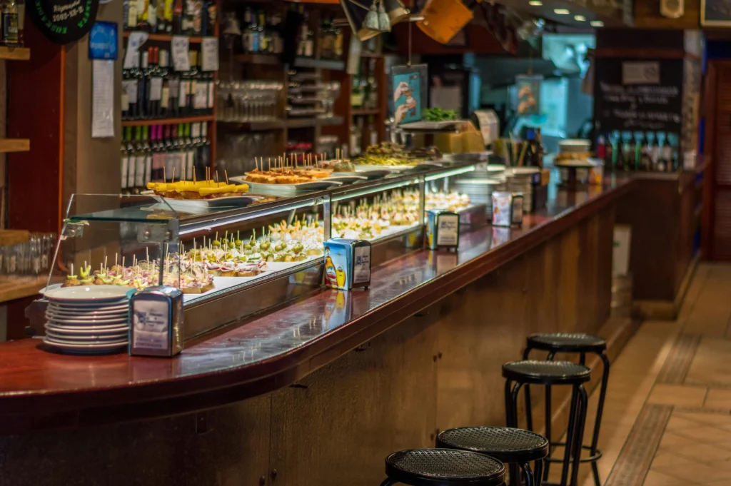 Vista de una barra tradicional en un bar de Barcelona, llena de tapas y pinchos variados expuestos en vitrinas de cristal, con taburetes y ambiente acogedor.