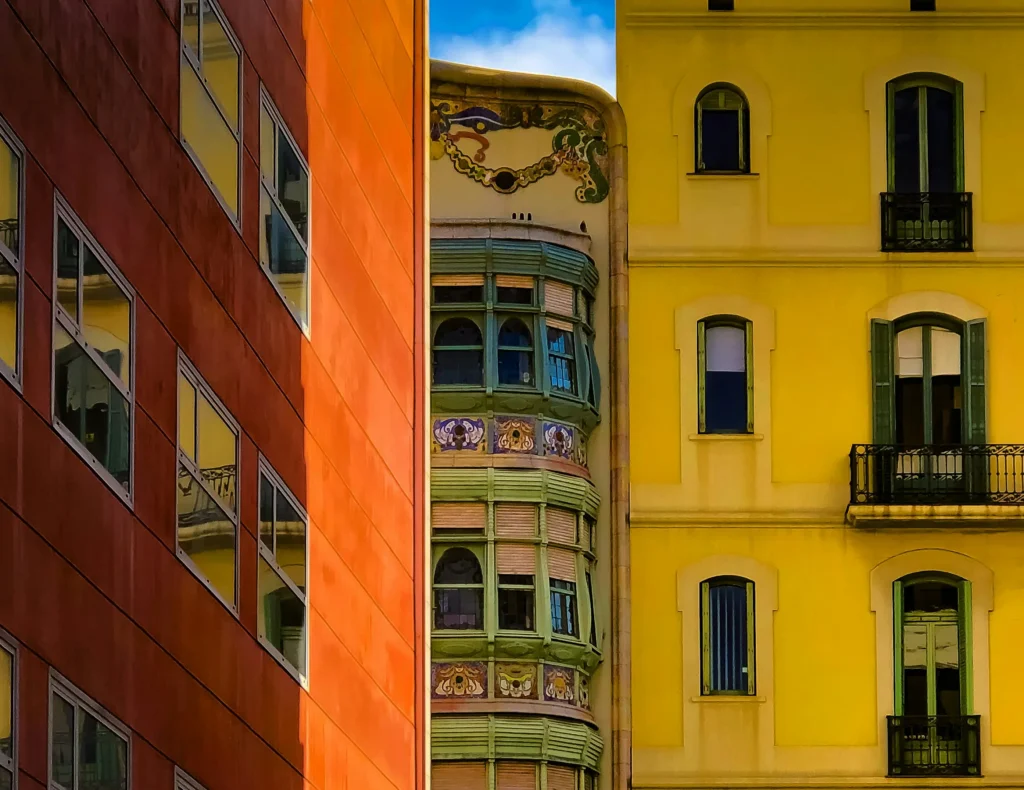 Vista de tres edificios en Barcelona que muestran un contraste arquitectónico: una fachada moderna de color rojizo con ventanas de cristal, un edificio amarillo clásico con balcones de hierro forjado y, en el centro, una construcción modernista con detalles decorativos y balcones verdes.