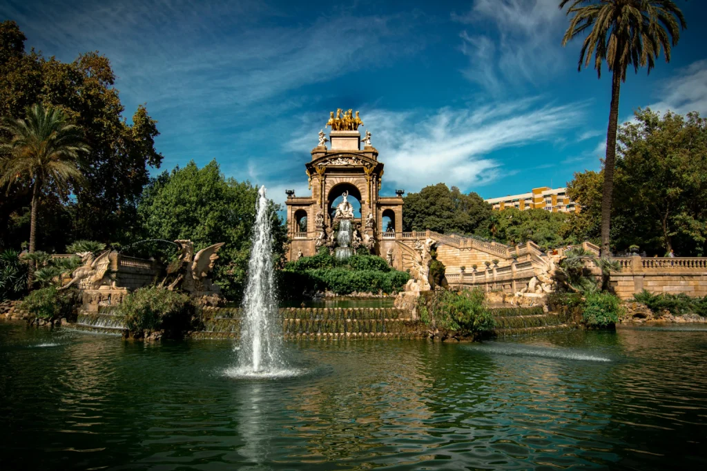 Vista frontal de la fuente monumental del Parque de la Ciutadella en Barcelona, con esculturas, cascadas y un chorro de agua en el estanque bajo un cielo azul.