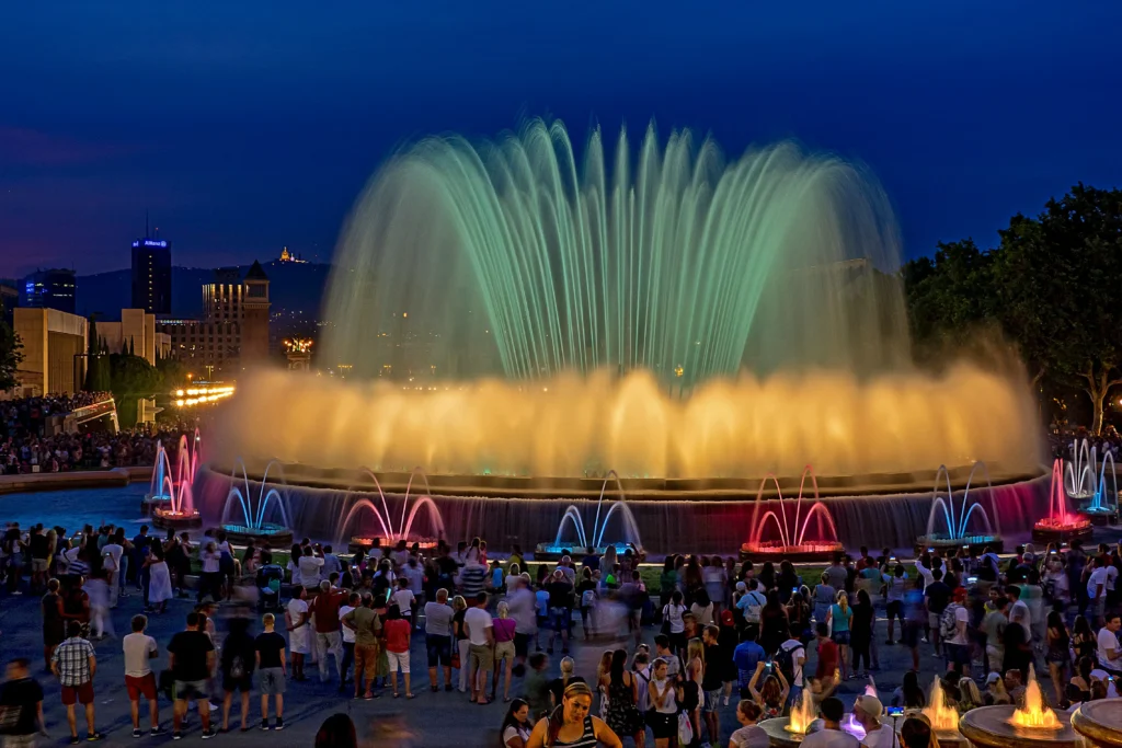 Espectáculo nocturno de luces y agua en la Fuente Mágica de Montjuïc en Barcelona, con una multitud de personas disfrutando del show frente a los chorros iluminados en colores vibrantes.