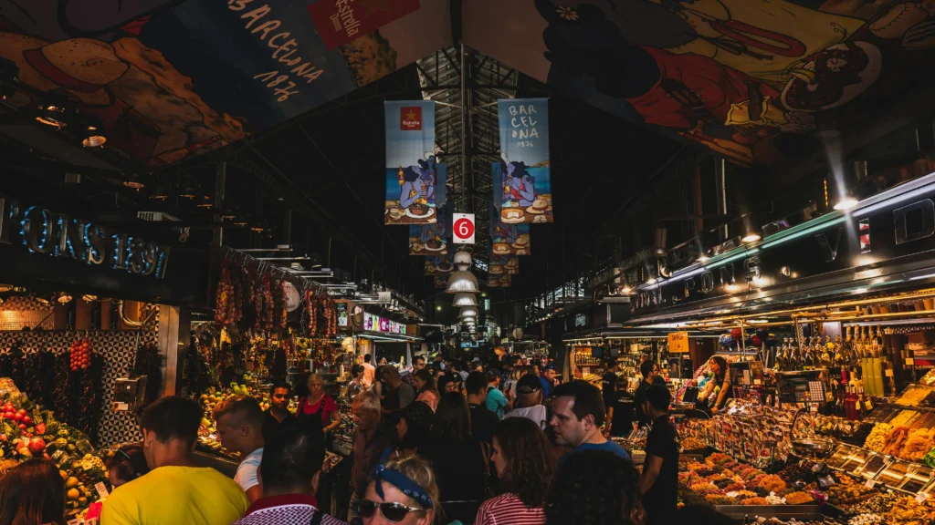 Vista del interior del Mercado de La Boqueria en Barcelona, lleno de puestos de frutas, embutidos y dulces, con una multitud de visitantes recorriendo los pasillos.