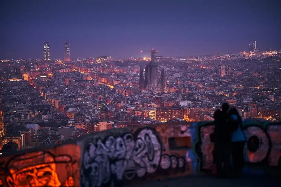 Panorámica nocturna de Barcelona desde los Búnkers del Carmel, con la Sagrada Familia iluminada en el centro y una pareja contemplando la ciudad en primer plano.