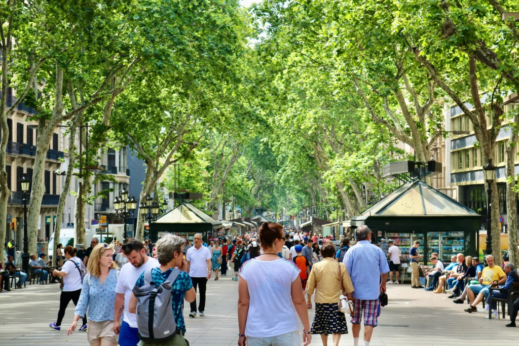 Vista de Las Ramblas en Barcelona con numerosos turistas paseando bajo los árboles y paradas de kioscos a ambos lados del paseo.
