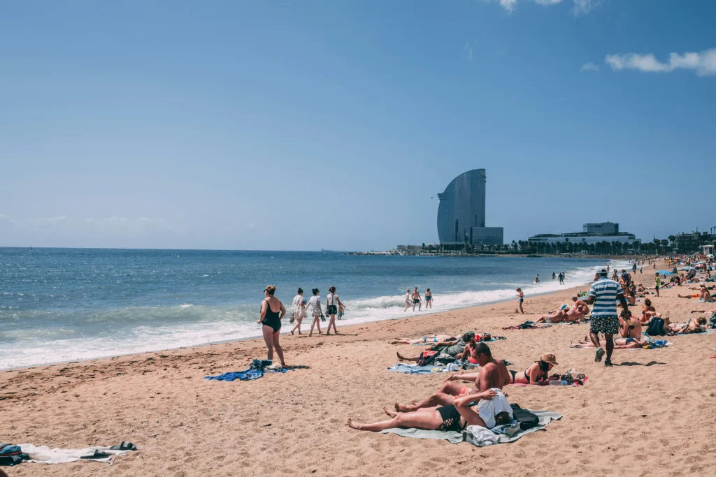 Personas disfrutando de un día soleado en la playa de la Barceloneta en Barcelona, con el mar Mediterráneo y el icónico Hotel W visible al fondo.