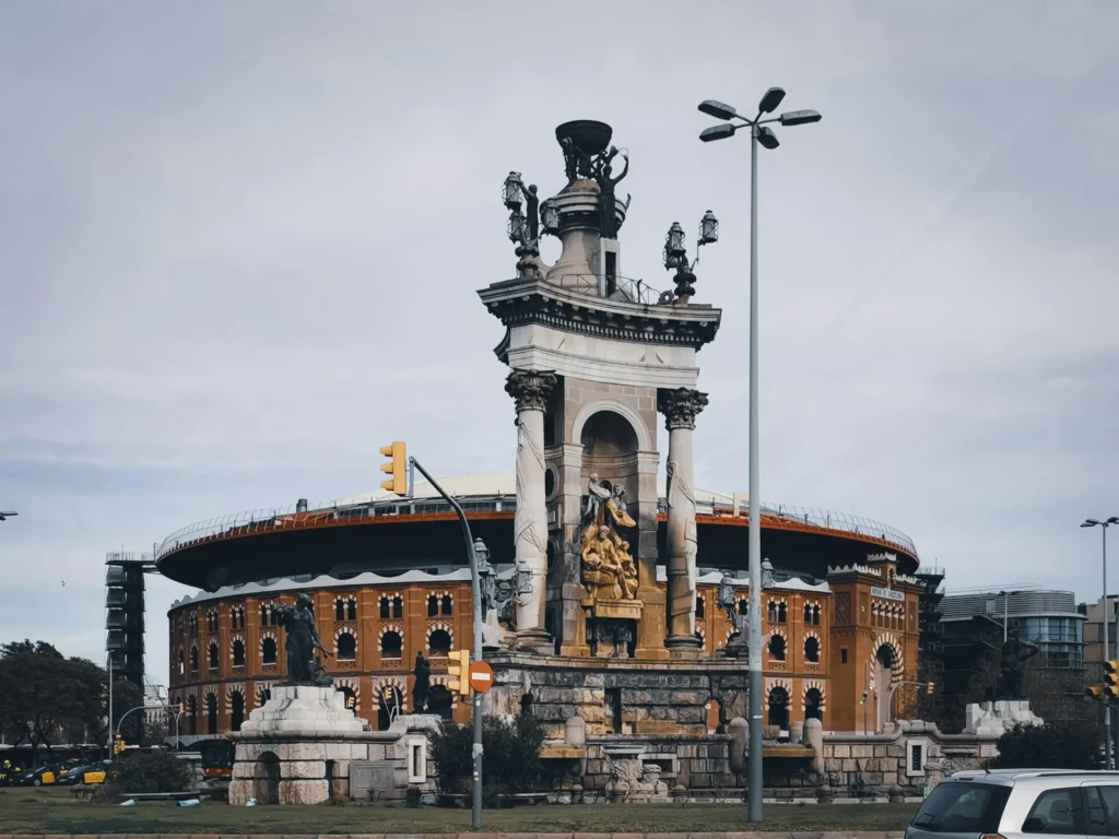 Vista del monumento central de la Plaza de España en Barcelona, con la antigua plaza de toros Las Arenas, hoy centro comercial, al fondo en un día nublado.