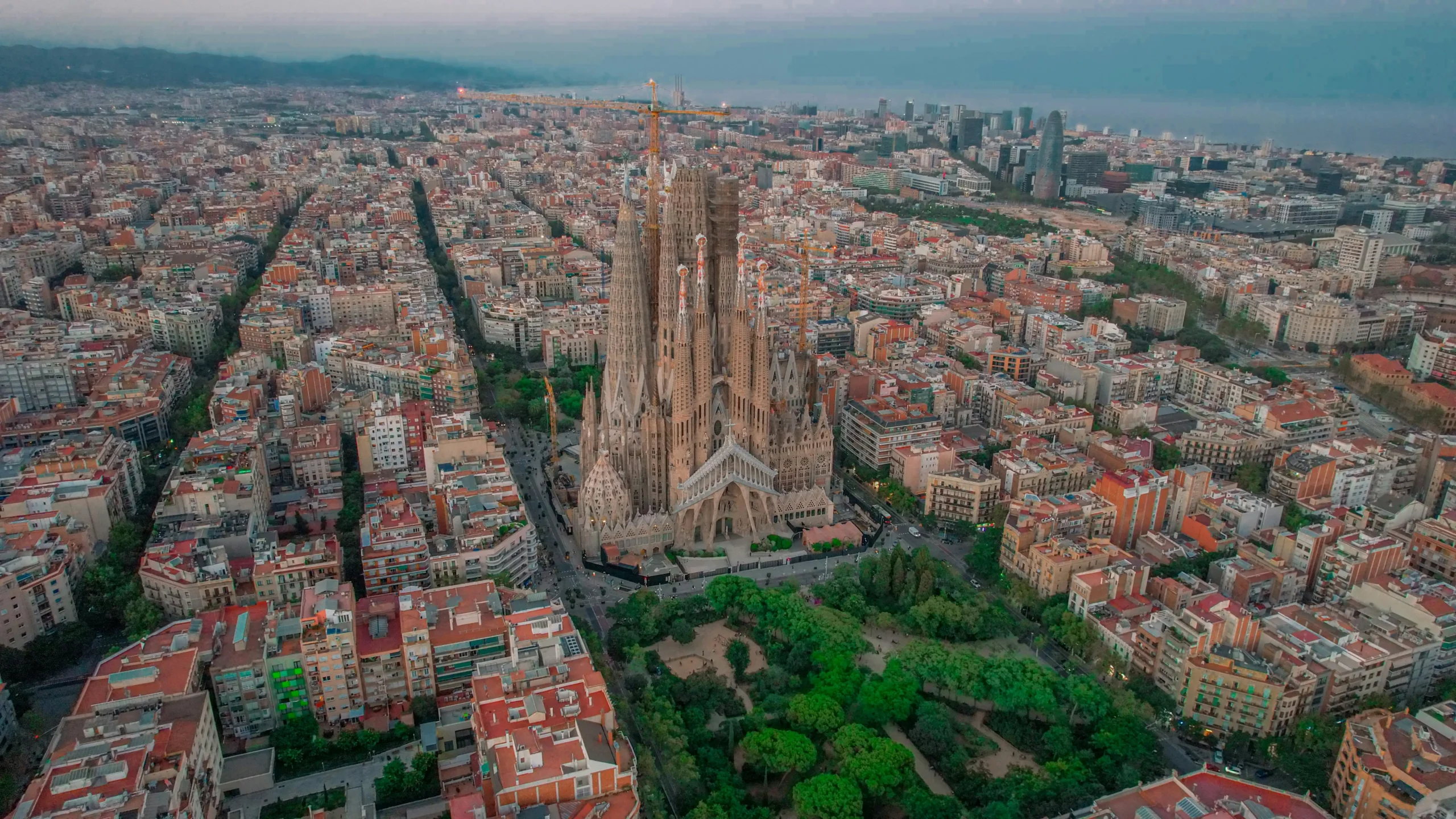 Aerial view of the Sagrada Familia in Barcelona, surrounded by the cityscape with green spaces and residential buildings, as seen during sunset.