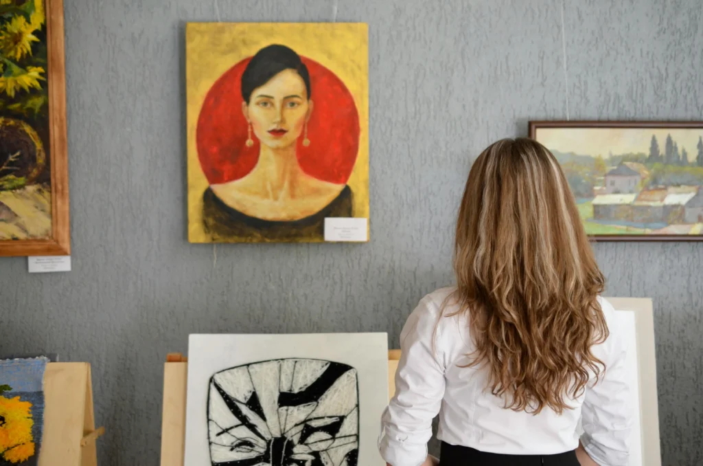 Woman with long wavy hair standing in front of a wall with various paintings at an art gallery, observing a portrait of a woman with a red circle background
