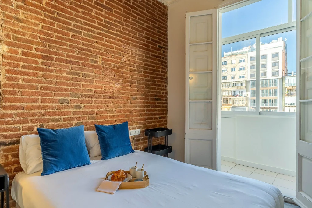 A stylish bedroom with an exposed brick wall, a neatly made bed with blue cushions, and a breakfast tray, next to a bright window leading to a balcony with city views