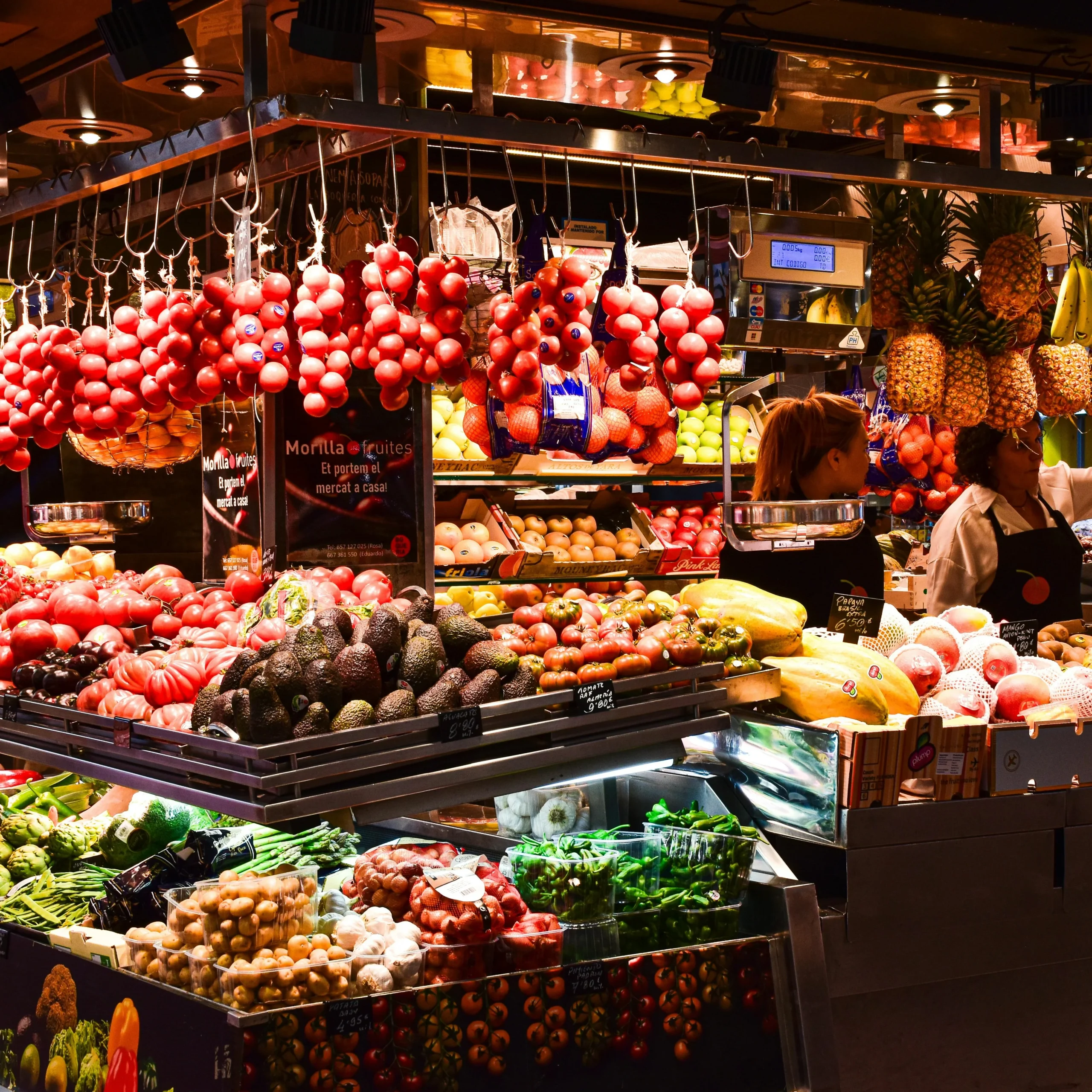 A vibrant fruit stand at a Barcelona market displaying fresh tomatoes, pineapples, bananas, and various exotic fruits.