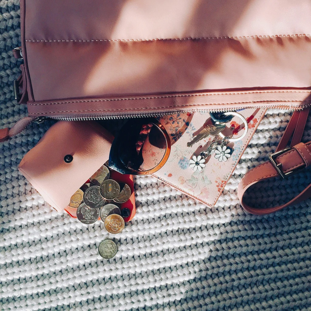 A pink handbag with sunglasses, coins, and accessories scattered on a textured surface, bathed in soft sunlight.