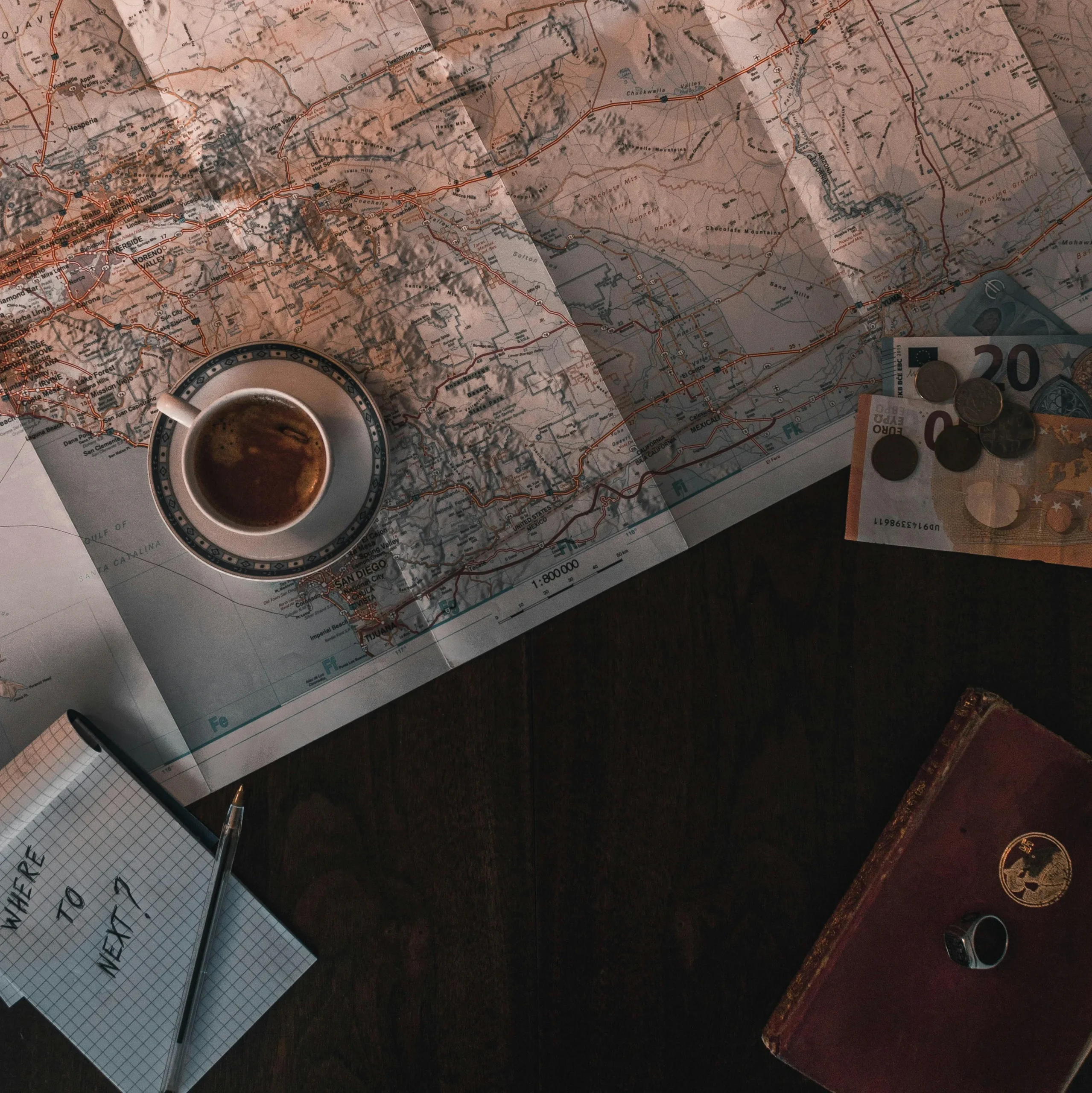 A vintage travel map laid out on a wooden table with a cup of coffee, money, and a notebook that says ‘Where to next?’