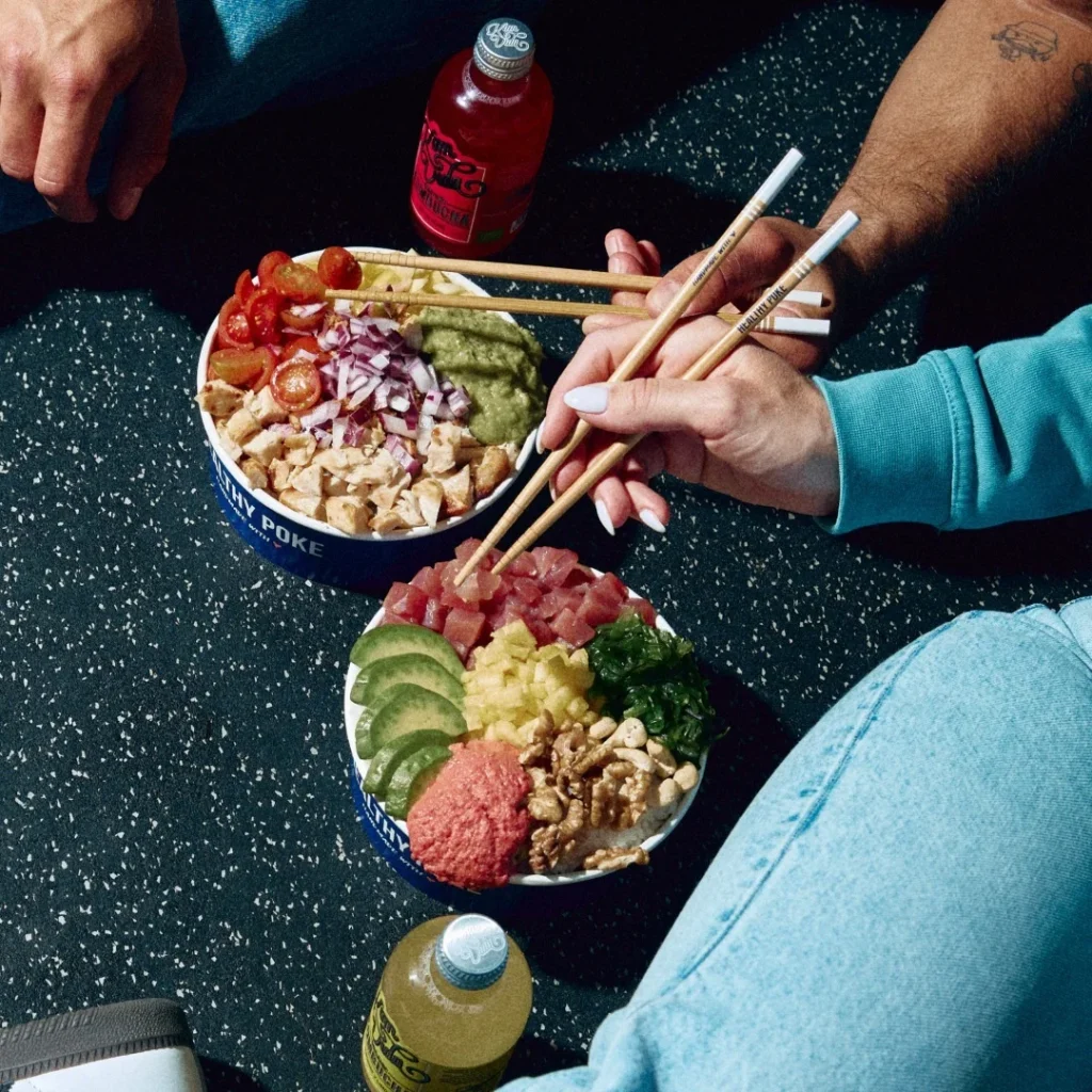 Two hands using chopsticks to share fresh poke bowls filled with tuna, avocado, cherry tomatoes, and crunchy nuts, enjoyed on a casual setting.