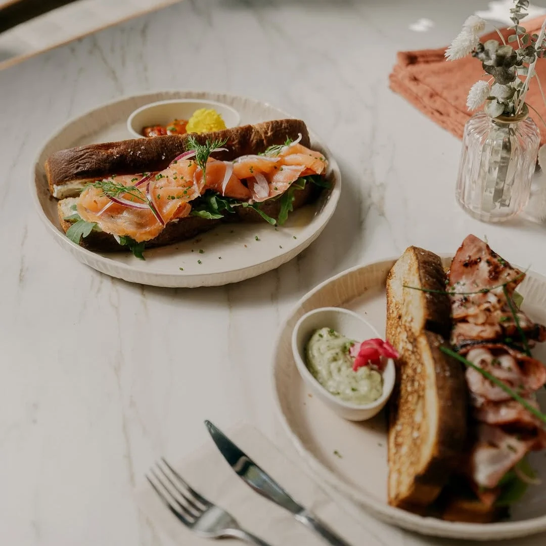 A beautifully plated smoked salmon sandwich on crusty bread, served with a side of avocado dip and a bacon sandwich on a marble table.