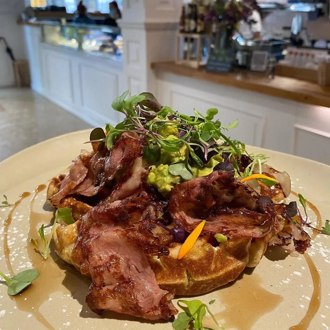 A golden waffle topped with crispy bacon, mashed avocado, and microgreens, drizzled with syrup, served in a cosy café.