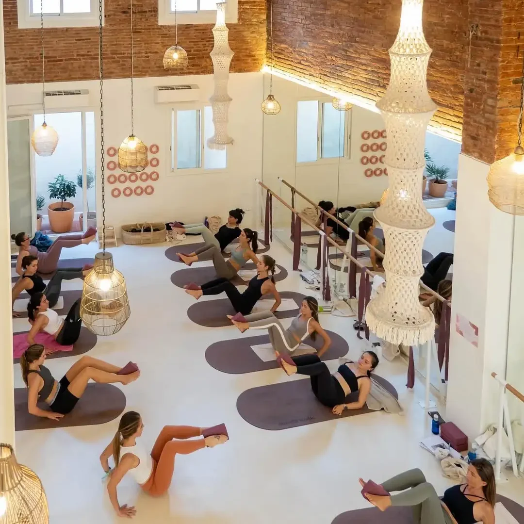 Group of women practicing yoga or Pilates in a bright, modern studio with exposed brick walls, large mirrors, and elegant hanging lights. Each participant is on a mat, using yoga blocks for support during a seated core exercise. The space features natural light from upper windows, plants in terracotta pots, and soft, minimalist decor.