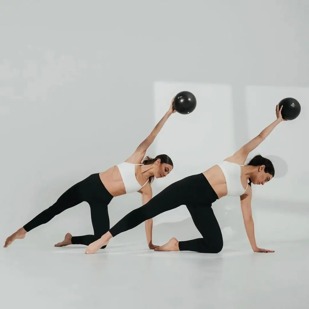 Two women performing a Pilates balance exercise in a bright, minimalist studio. Both are in a side kneeling position, wearing black leggings and white sports bras, holding black weighted balls with one arm extended upward. The clean white background and soft shadows create a calm, focused atmosphere.