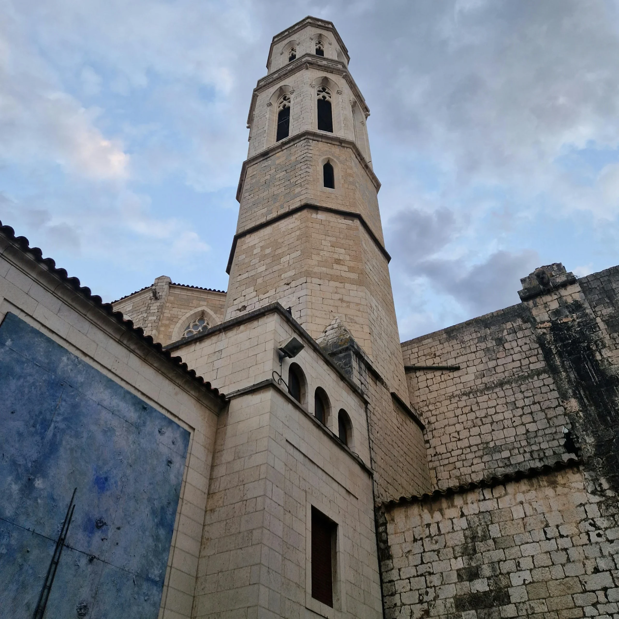 A towering bell tower of a historic church stretches into the sky, captured from below. The stone facade of the building is weathered with time, contrasting against the dramatic, cloudy sky. The scene evokes a sense of grandeur and history, with intricate architectural details visible in the tower and surrounding wall