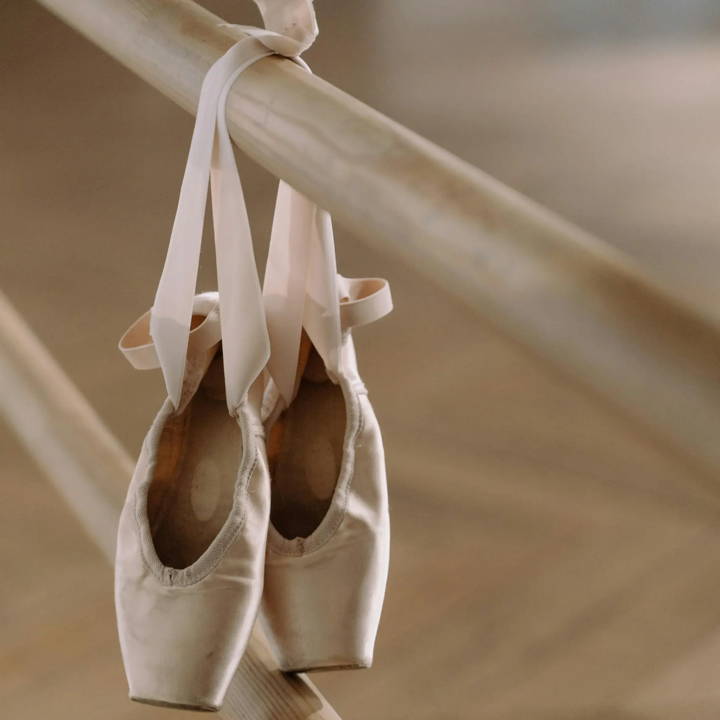 Close-up of a pair of worn pink ballet pointe shoes hanging by their ribbons on a wooden ballet barre in a softly lit dance studio. The background is blurred, emphasizing the delicate texture of the shoes and the serene atmosphere of the space.