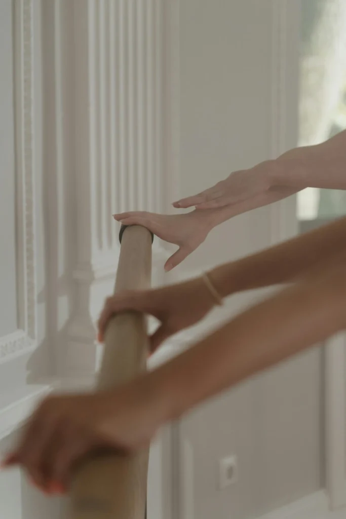 Close-up of two people gently placing their hands on a wooden ballet barre in a bright, softly lit studio with classic white wall moldings. The image captures a calm, poised moment before or during a dance or barre workout session.