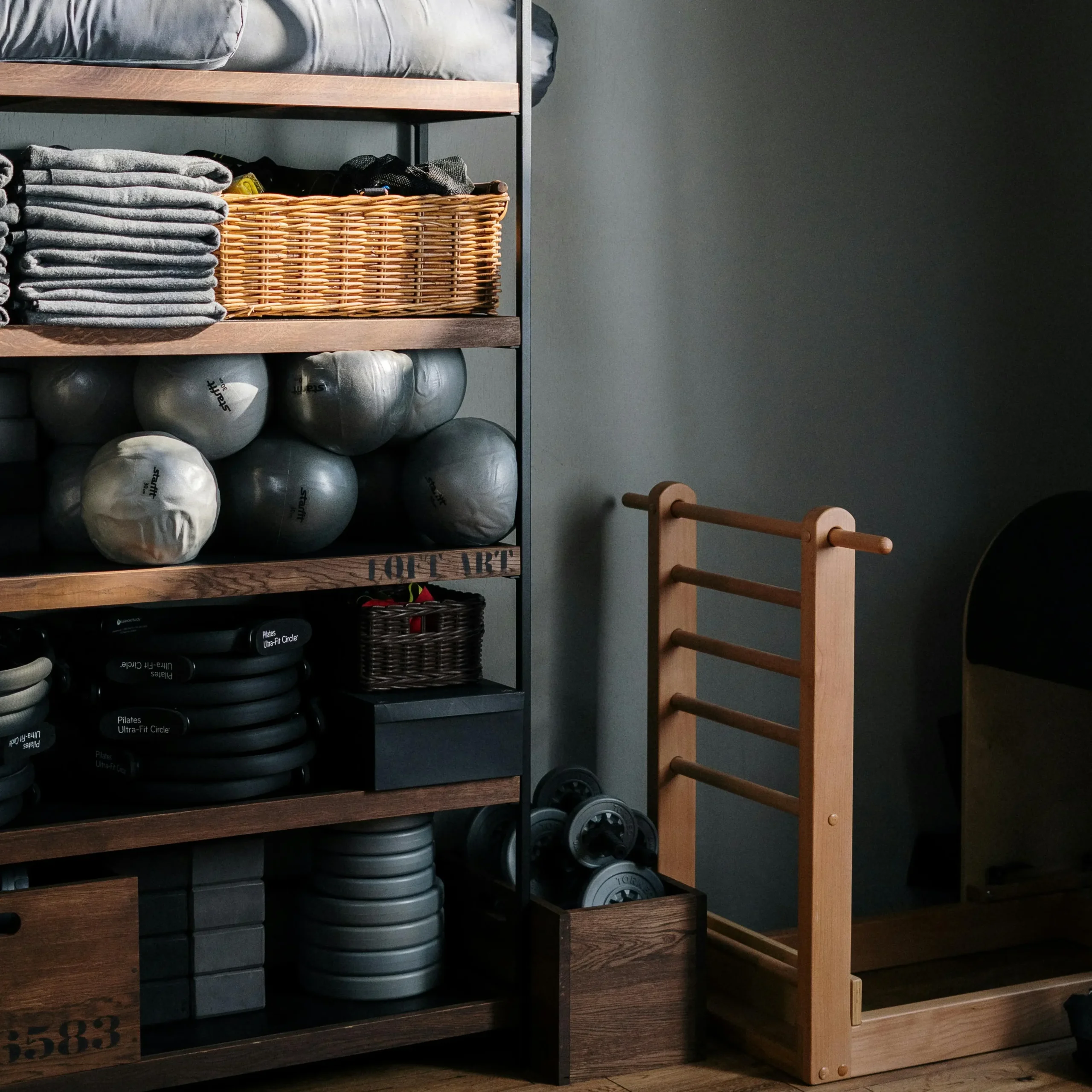 Neatly arranged shelves in a wellness or Pilates studio holding gray bolsters, folded towels, wicker baskets, fitness balls, resistance rings, and weights. A wooden ladder barrel and gym flooring complete the cosy, minimalist setup, bathed in soft natural light from the side.