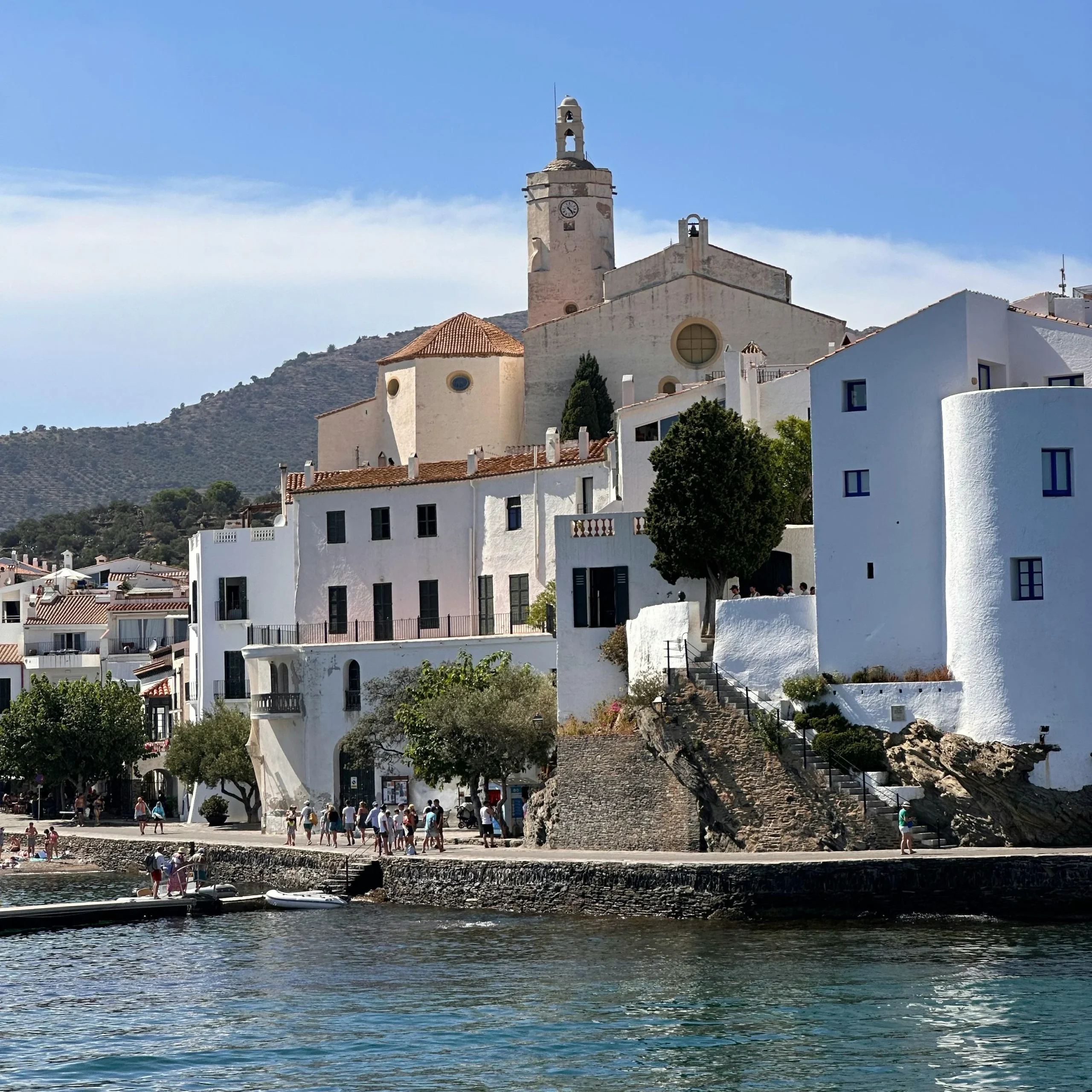 A beautiful view of the coastal town of Cadaqués, with its whitewashed buildings and the iconic clock tower of the Church of Santa Maria towering over the picturesque waterfront. The crystal-clear water of the Mediterranean Sea reflects the serene surroundings, while visitors stroll along the promenade beneath a bright blue sky