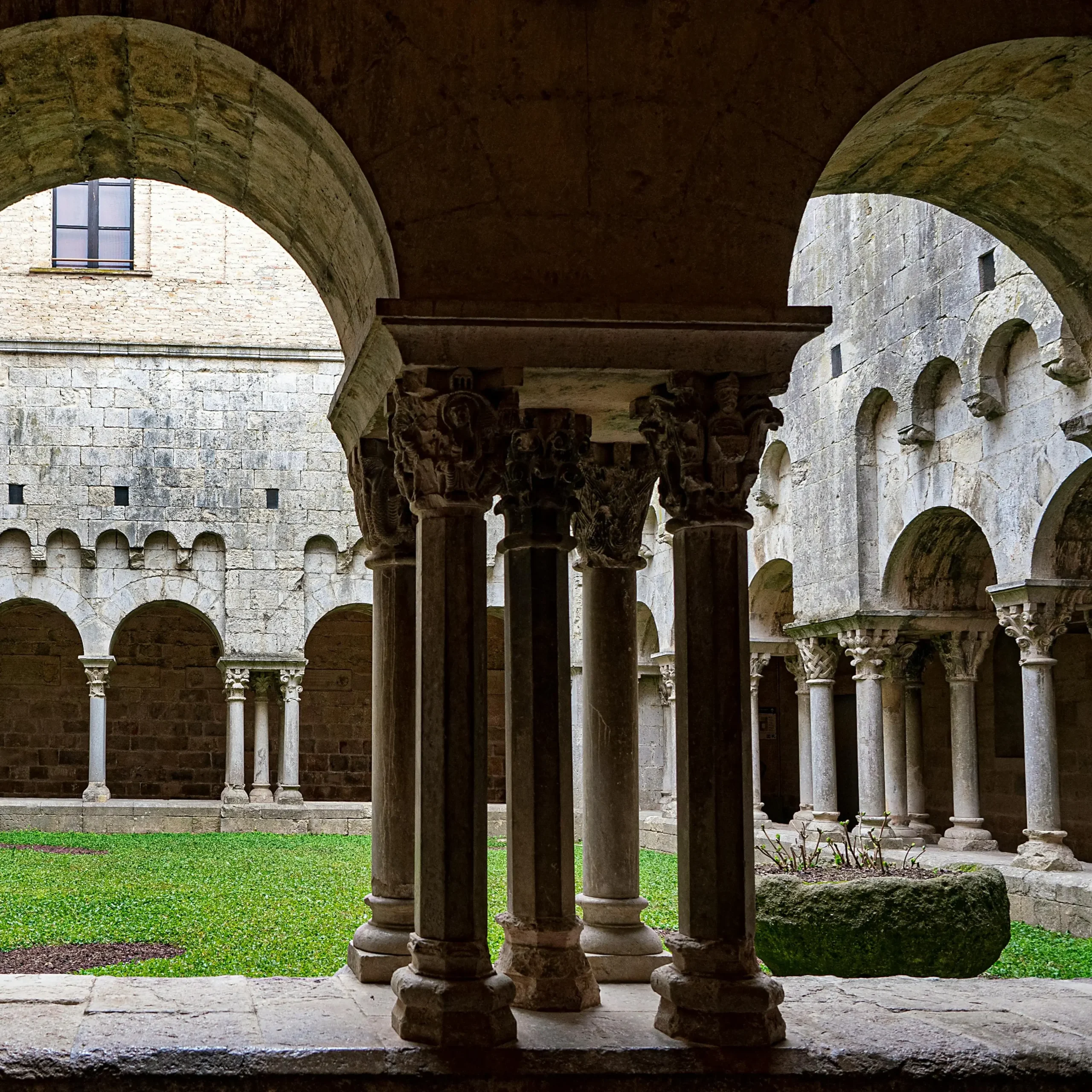 Romanesque cloister with stone arches and intricately carved columns surrounding a central grassy courtyard in a historic monastery or church.