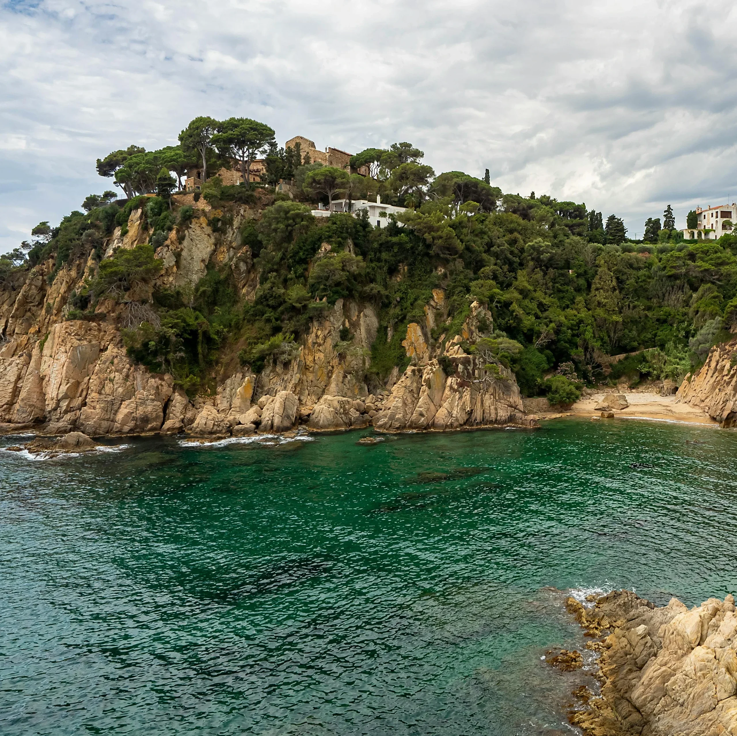 A picturesque view of a tranquil cove along the Costa Brava, with clear turquoise waters gently lapping against the rocky shoreline. Towering cliffs adorned with lush greenery rise above, with elegant villas nestled along the edge, offering an idyllic coastal escape