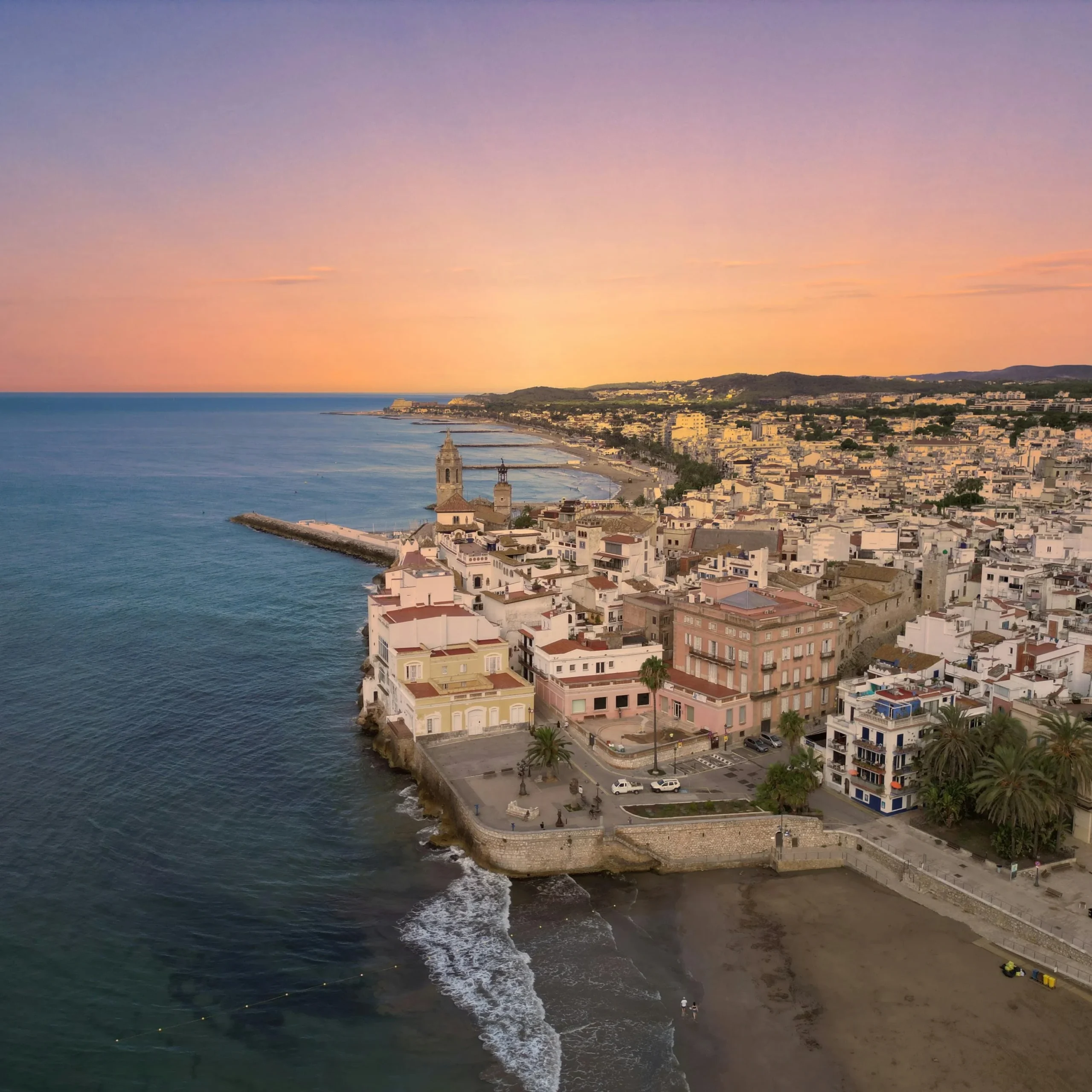 A stunning aerial view of Sitges, a coastal town in Catalonia, Spain, captured during sunset. The golden light reflects off the buildings and the Mediterranean Sea, with the sandy beach stretching into the distance. The town’s charming white buildings, palm trees, and iconic church tower add to the picturesque landscape.