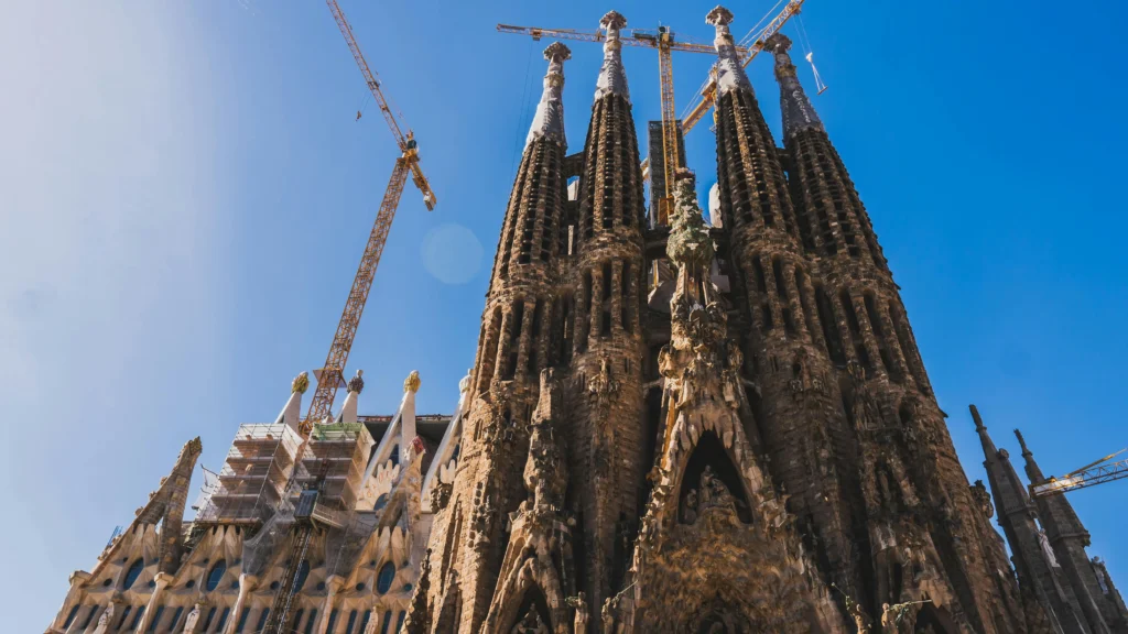 Close-up view of the Sagrada Família basilica in Barcelona with cranes above, highlighting its intricate towers and ongoing construction against a clear blue sky.