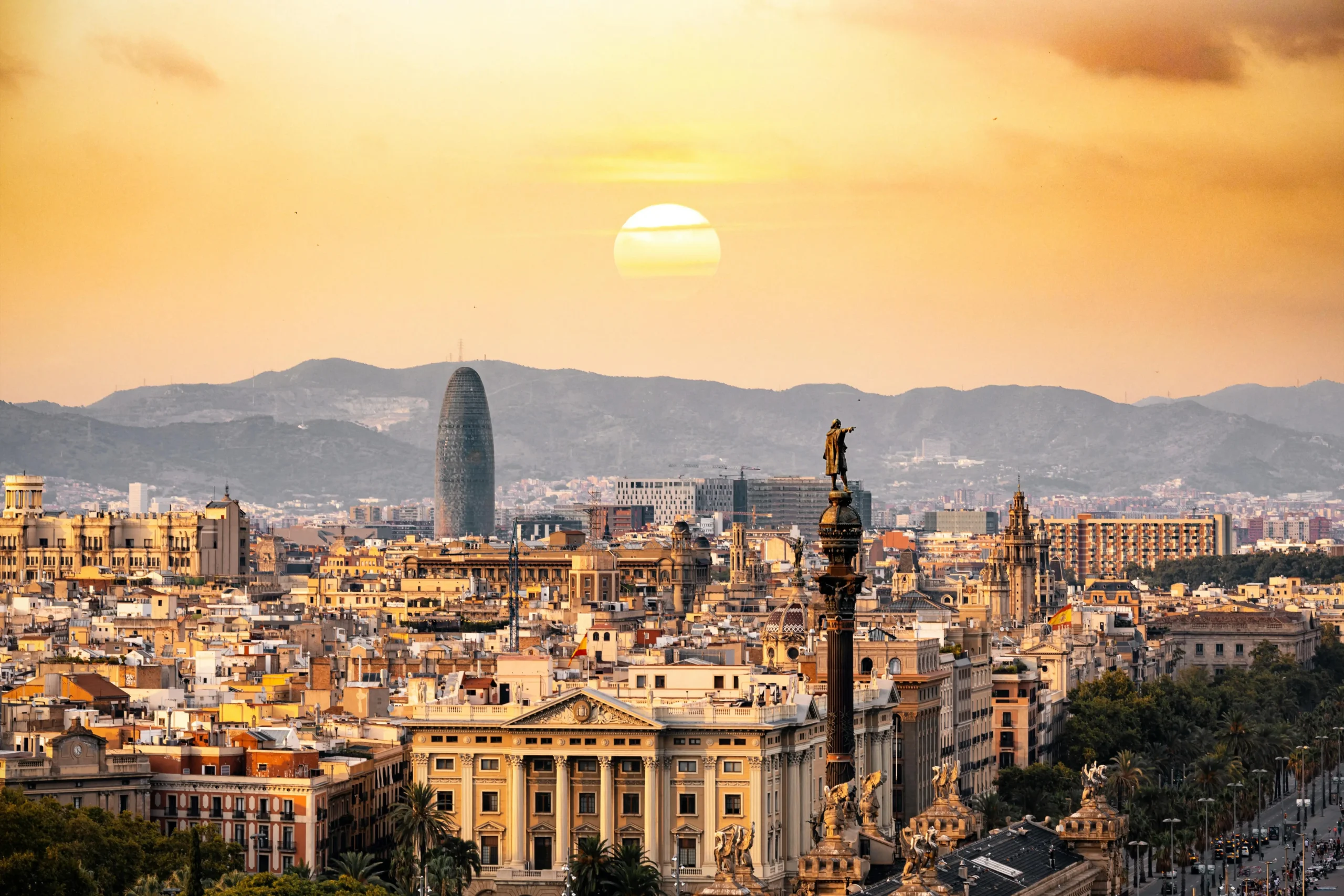 A breathtaking view of Barcelona's skyline at sunset, with landmarks such as the Columbus Monument and the Torre Agbar standing tall against the vibrant colors of the sky.