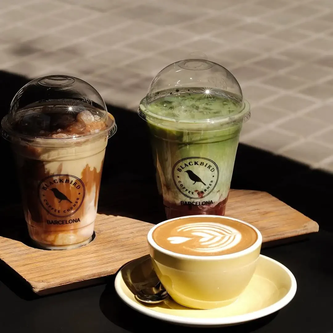 Zwei Eiskaffees und ein Flat White von Blackbird Coffee Corner in Barcelona, darunter ein Matcha Latte, ein Karamell-Eiskaffee und ein Cappuccino mit Latte Art, serviert auf einer sonnigen Terrasse.