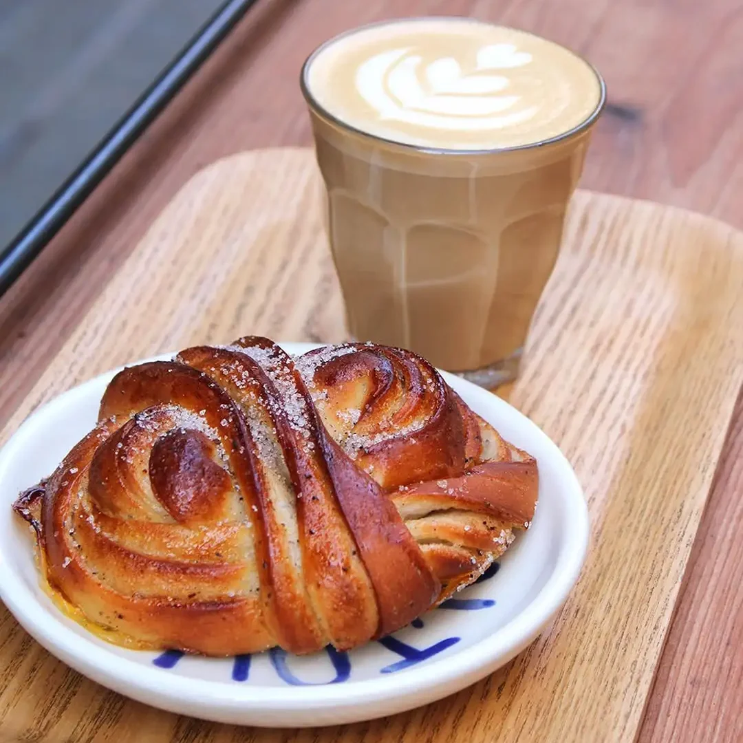 Latte mit Latte Art und Kardamomschnecke von Origo Bakery in Barcelona, serviert auf einem Holztablett an einem Außentisch.