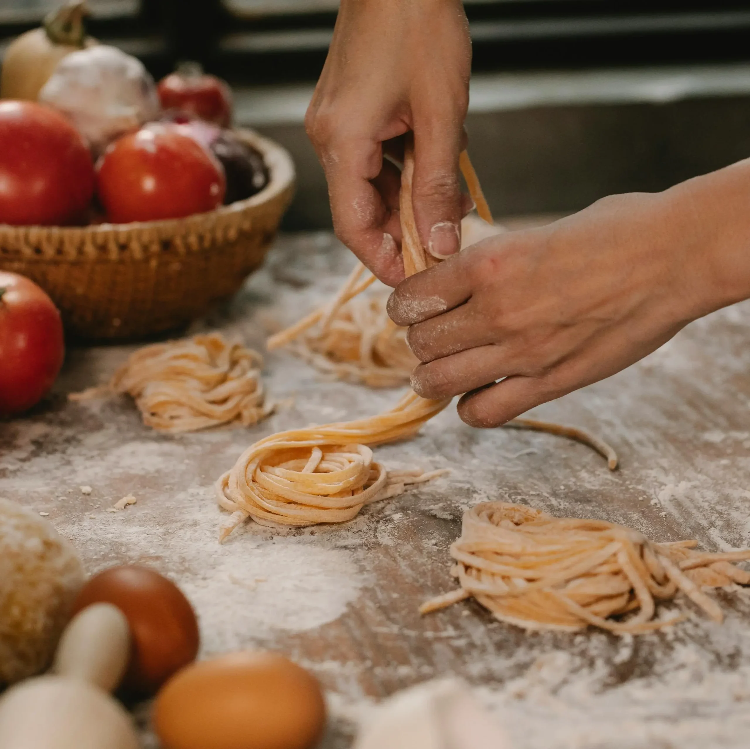 Hands shaping fresh homemade pasta nests on a floured wooden surface with tomatoes, eggs, and dough nearby.