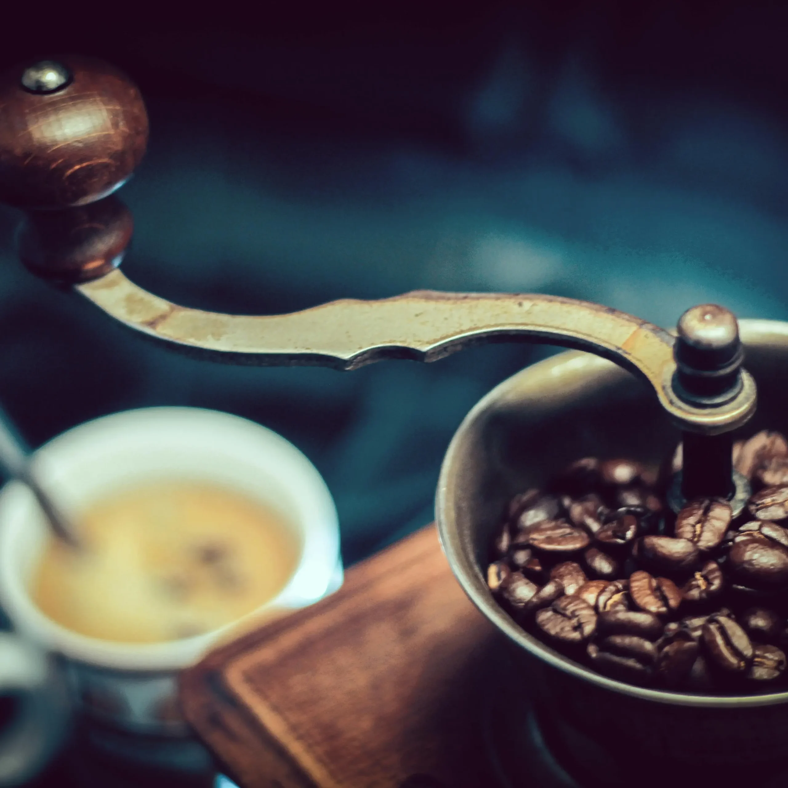 Vintage-Handkaffeemühle mit gerösteten Bohnen, im Hintergrund ein unscharfer Espresso.