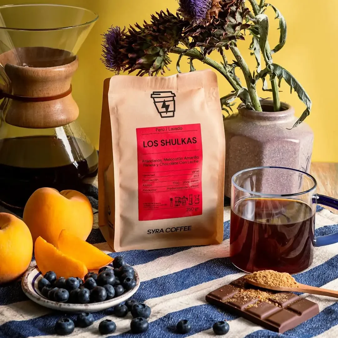 Spezialitätenkaffeebohnen von Syra Coffee in Barcelona mit der Mischung 'Los Shulkas' aus Peru. Geschmacksnoten: Blaubeere, gelber Pfirsich, Panela und Milchschokolade, präsentiert mit frischem Obst und einem Chemex.