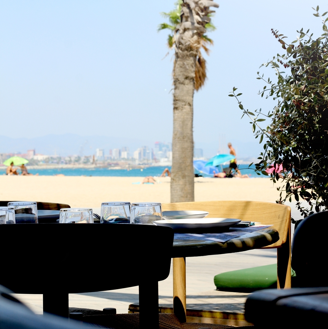 Outdoor restaurant table set with plates and glasses, overlooking a sandy beach, palm trees, and the Mediterranean Sea on a sunny day.