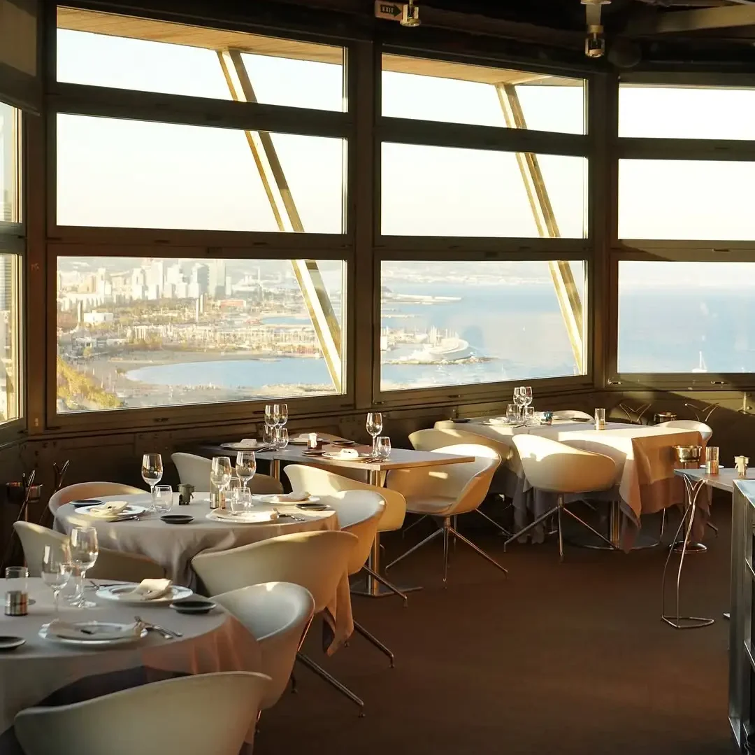 Elegant indoor restaurant with panoramic windows offering breathtaking views of the Barcelona coastline and Mediterranean Sea, tables set for fine dining.