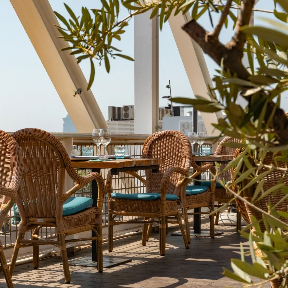 Stylish outdoor restaurant terrace with wicker chairs, turquoise cushions, and neatly set tables, partially shaded and overlooking the city and sea.