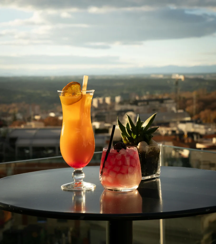 Two vibrant cocktails, one orange and one pink, served on a rooftop table with a city view in the background under a partly cloudy sky.