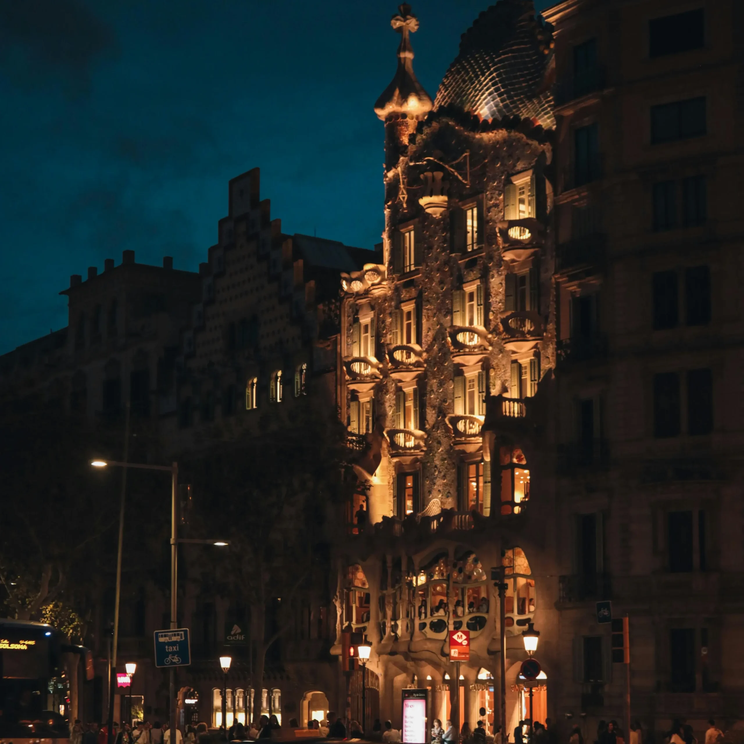 Night view of Casa Batlló in Barcelona, illuminated with warm lights, with a taxi passing by in the foreground.
