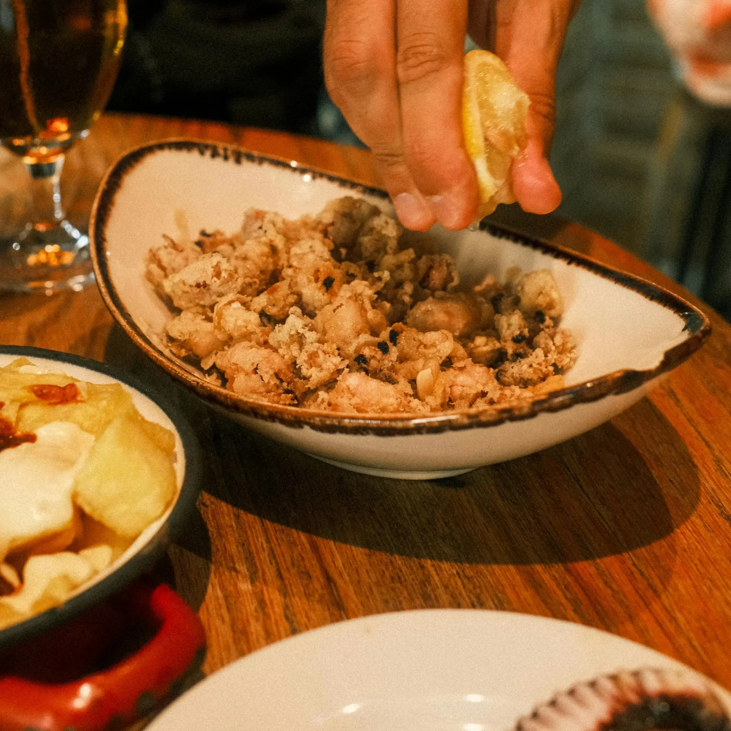 Close-up of a hand squeezing lemon over a bowl of crispy fried calamari, with a glass of beer and a side dish of potatoes in the background.