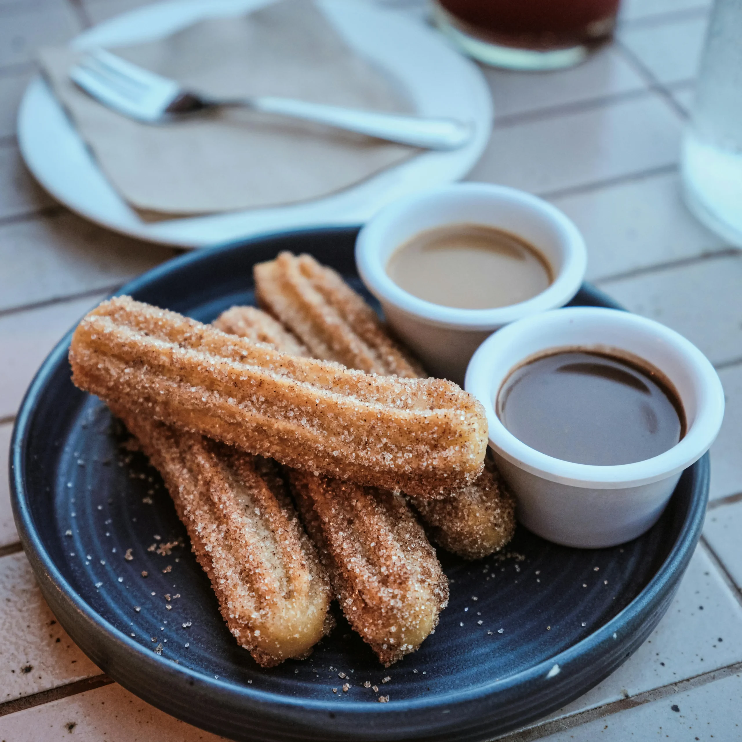 Plate of sugar-coated churros served with cups of chocolate and caramel dipping sauces, placed on a tiled table with drinks in the background.