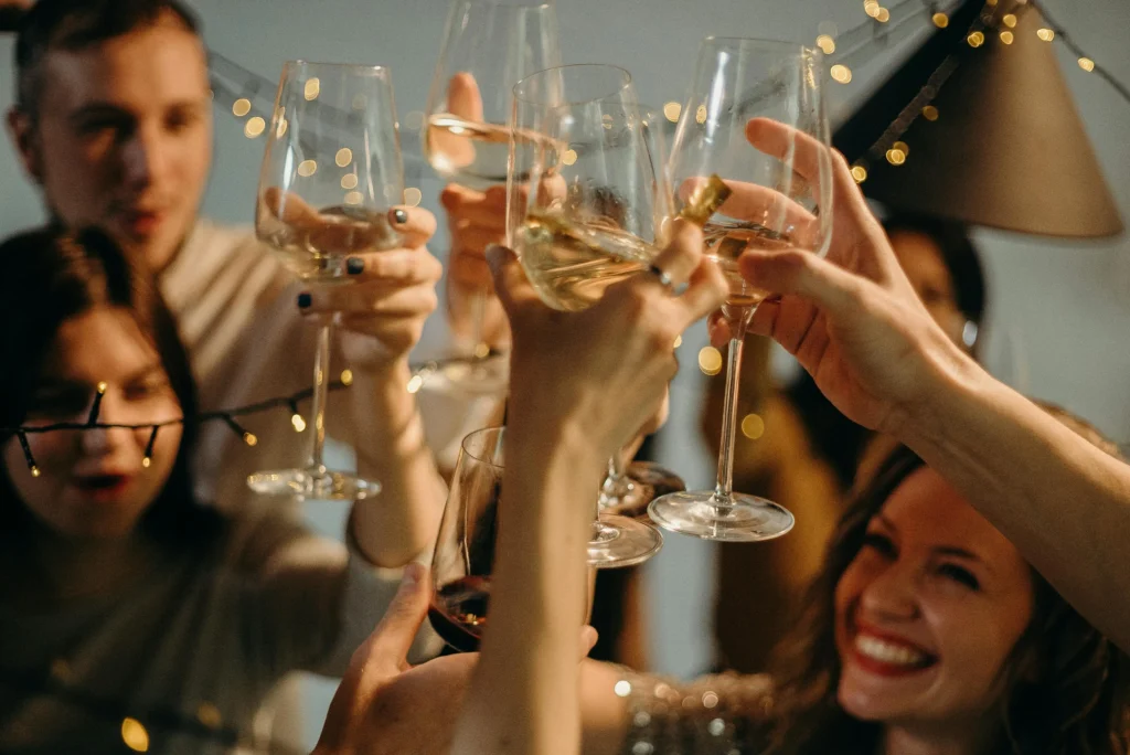 Close-up of friends raising wine glasses in a toast, with warm string lights in the background and a smiling woman in the foreground.