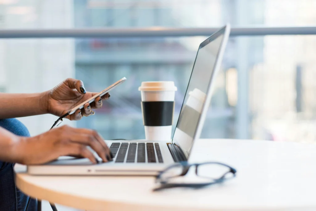 Person using a laptop and holding a smartphone at a desk with a coffee cup and glasses.