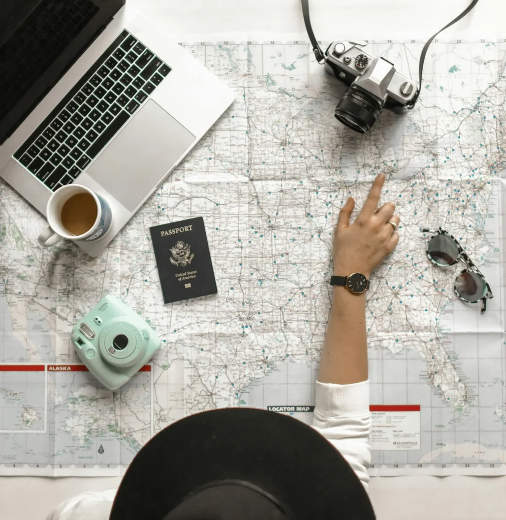 Person planning a trip with a map, pointing at a location, surrounded by a passport, coffee, camera, instant camera, sunglasses, and laptop.