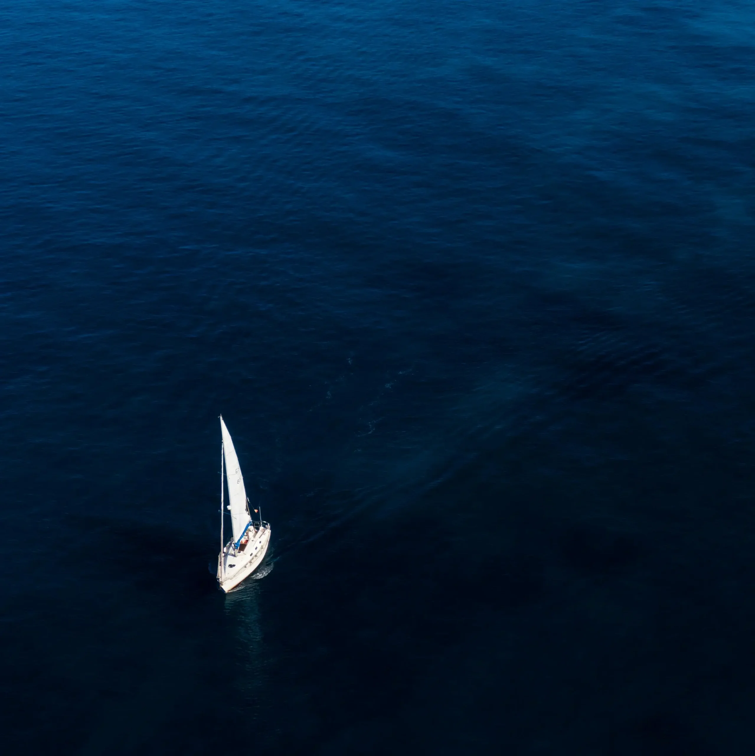 A white sailboat sailing alone across the deep blue ocean, photographed from above, surrounded by calm and vast open water.