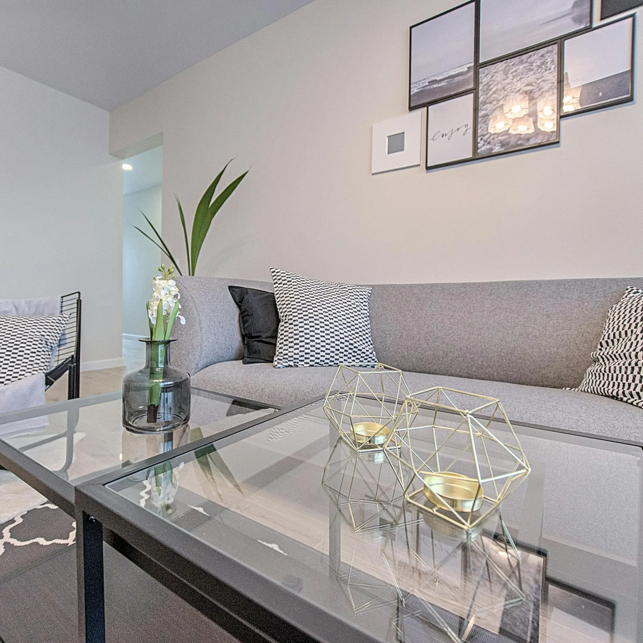 Stylish living room featuring a gray sofa with patterned cushions, a glass coffee table with geometric candle holders and a vase with flowers, and contemporary wall art.