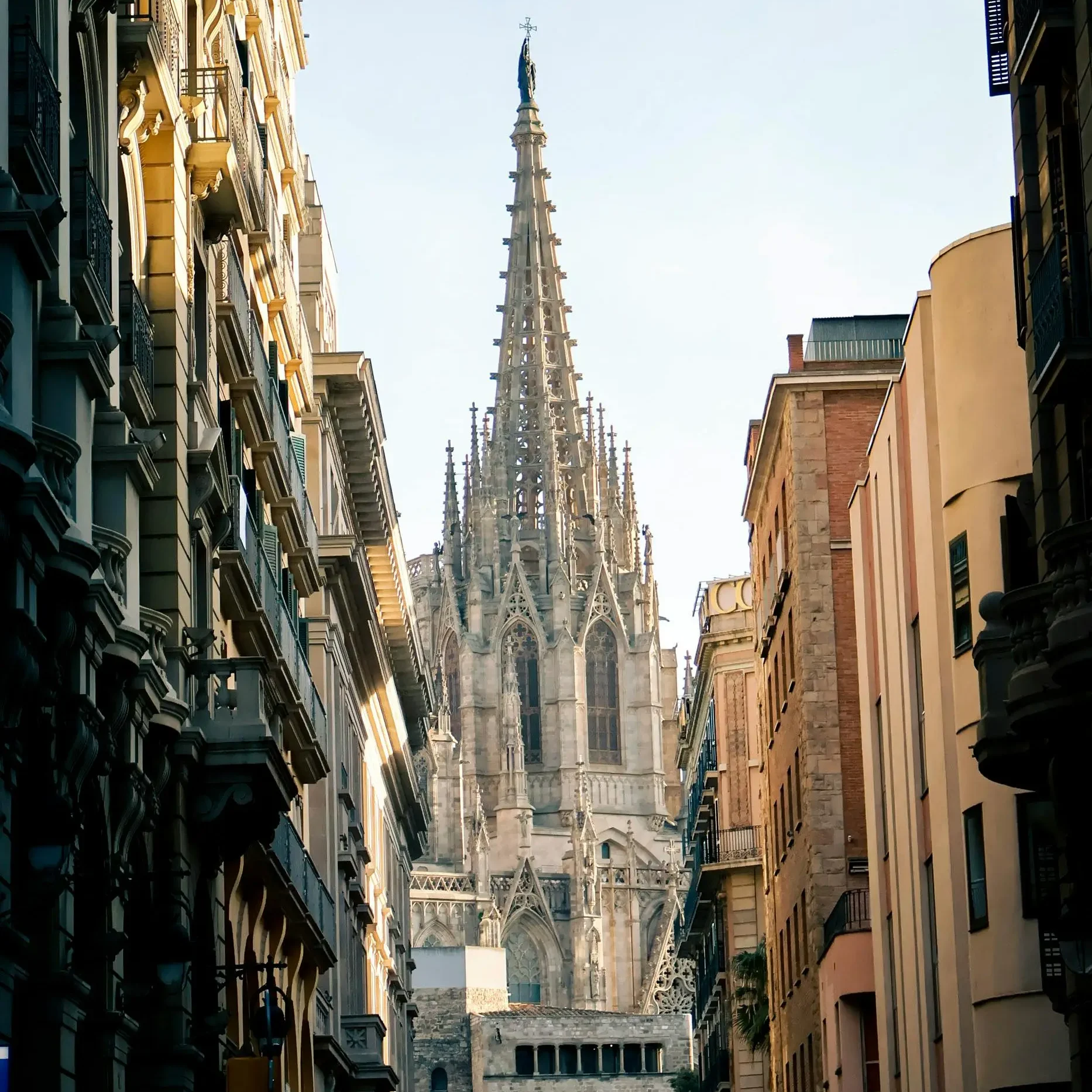 View of the Gothic spire of Barcelona Cathedral framed by narrow streets with historic buildings, under a clear blue sky.