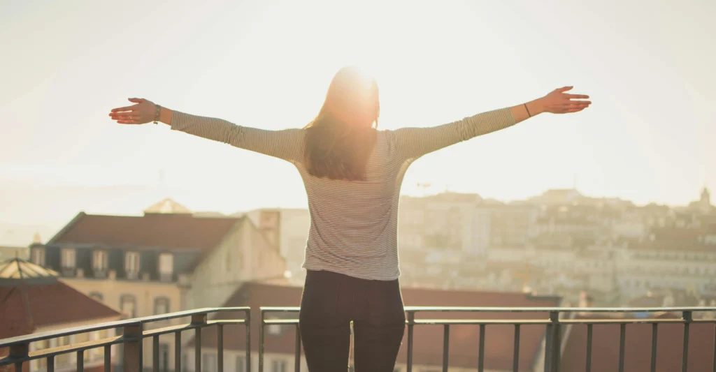 Woman standing on a balcony with arms outstretched, facing the sunrise over a cityscape.